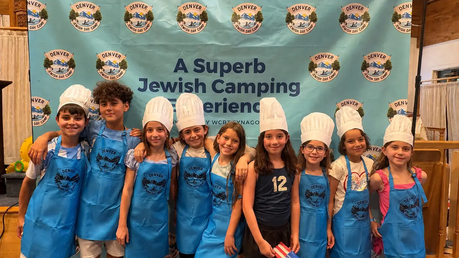 Campers in blue camp aprons and chef hats in front of the “A Superb Jewish Camping Experience” banner