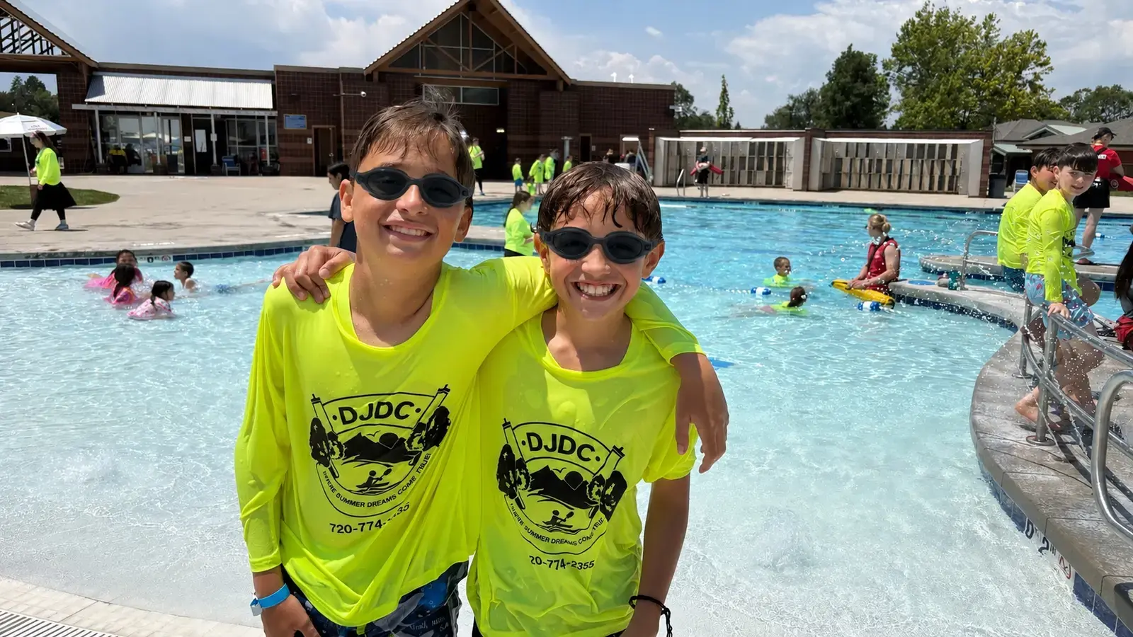 Two campers in neon DJDC shirts and sunglasses, arms around each other at the pool
